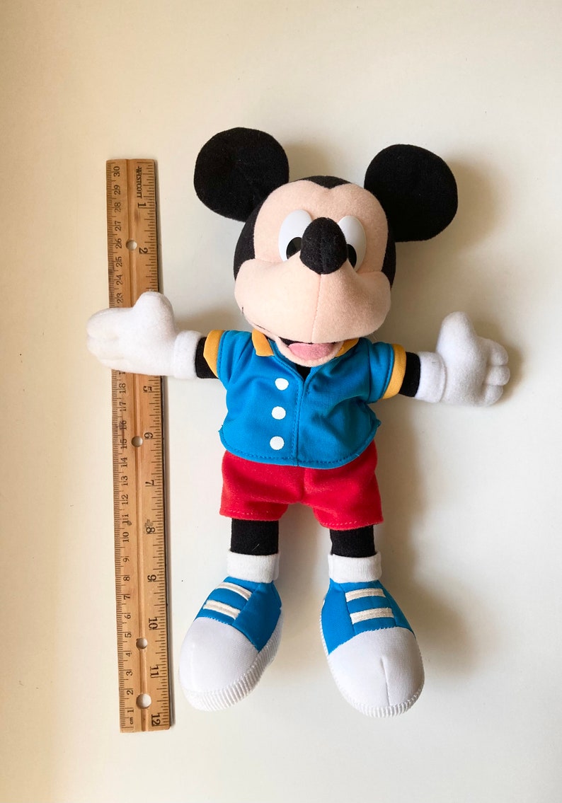 May include: A Mickey Mouse plush toy, wearing a blue jacket, red shorts, and blue shoes. The toy is approximately 12 inches tall, with a ruler placed next to it for scale. The classic character has black ears and white gloves.
