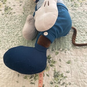 May include: A blue and brown plush toy of a cartoon character with large white gloves and a blue button on the pants. The toy is lying on a white and green floral patterned fabric with a measuring tape next to it.