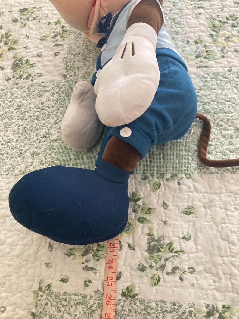 May include: A blue and brown plush toy of a cartoon character with large white gloves and a blue button on the pants. The toy is lying on a white and green floral patterned fabric with a measuring tape next to it.