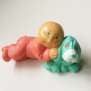 May include: A plastic figurine of a baby doll in a pink sleeper hugging a turquoise dog figurine.