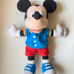 May include: A plush Mickey Mouse doll, featuring black ears, a black face, and a tan muzzle. He wears a blue shirt with yellow trim, red shorts, and blue shoes with white soles. The doll's arms are outstretched.