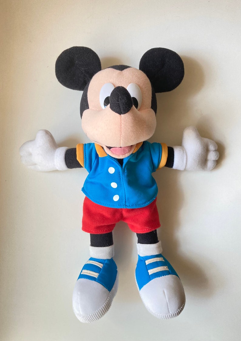 May include: A plush Mickey Mouse doll, featuring black ears, a black face, and a tan muzzle. He wears a blue shirt with yellow trim, red shorts, and blue shoes with white soles. The doll's arms are outstretched.