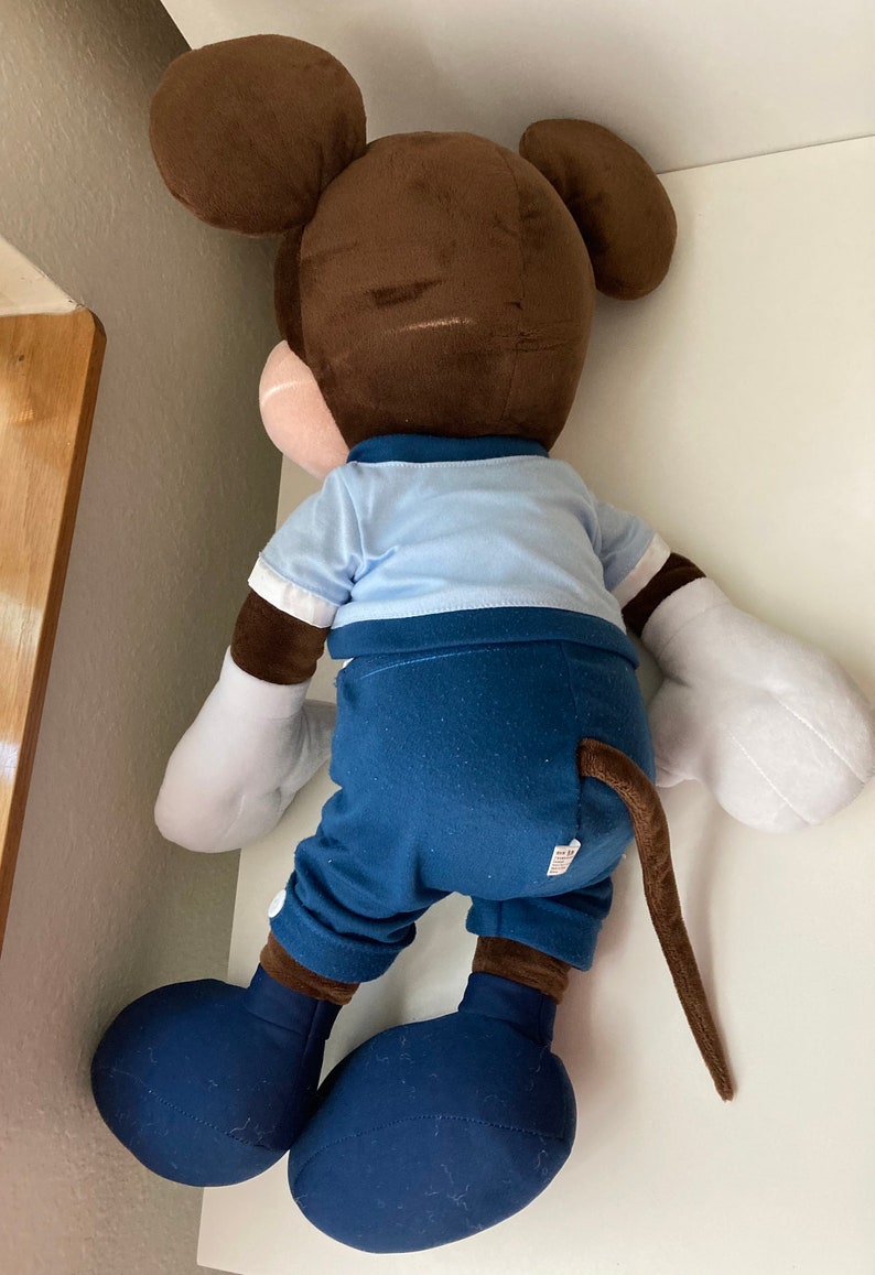 May include: A large brown and blue plush Mickey Mouse toy. The toy is wearing a blue shirt and blue pants. The toy has a long brown tail.