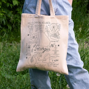 Può includere: Una borsa tote beige con un'illustrazione in bianco e nero dei monumenti della Georgia. Il design include il contorno dello stato, le parole "Georgia", "Atlanta", "Pecan Pie", "Peach State" e altre icone.