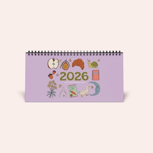 Puede incluir: Calendario de escritorio de 2026 con espiral y fondo lila. Presenta ilustraciones de una manzana, un cruasán y un caracol. El texto dice: «A lot can happen in a year». Diseñado para uso en escritorio.