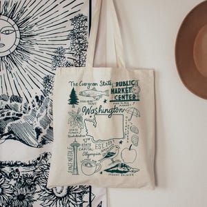 Può includere: Borsa tote color crema con una mappa illustrata in verde dello stato di Washington. Il design include la Space Needle, il Monte Rainier e il testo "The Evergreen State Public Market Center". La borsa è appesa a una parete bianca.