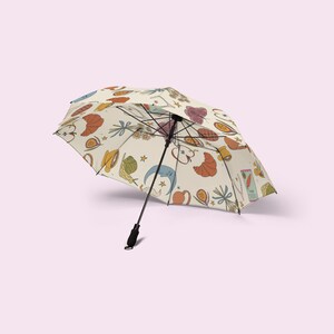 May include: An open umbrella with a cream-colored canopy featuring a repeating pattern of colorful illustrations. The illustrations include croissants, fruit, and other food items. The umbrella has a black handle and frame.