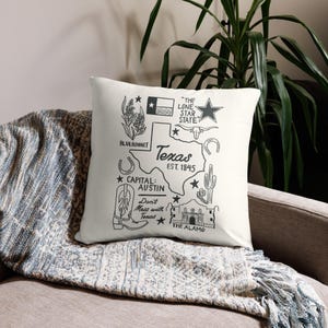 Peut inclure: Coussin carré blanc cassé avec un motif noir sur le thème du Texas. Le motif comprend le contour de l'État, le texte "Texas EST. 1845" et des illustrations d'un lupin, d'une botte de cowboy et de l'Alamo. Le coussin est sur une couverture grise et bleue.