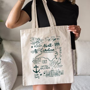 Può includere: Una borsa tote beige con un'illustrazione verde dei punti di riferimento della Carolina del Nord, tra cui un aereo, un'ape e il Biltmore Estate. Il testo include "North Carolina" e "EST 1789."