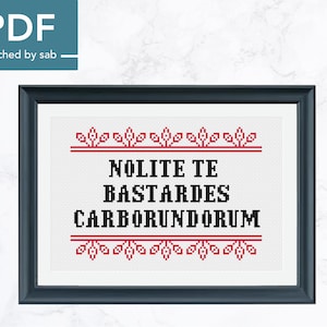 May include: A black and white cross-stitch pattern with the Latin phrase "Nolite Te Bastardes Carborundum" in a black font. The phrase is surrounded by a red border with small red flowers.