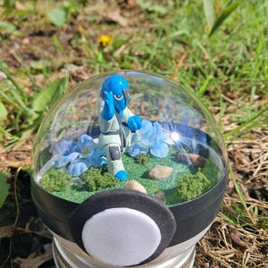 May include: A terrarium featuring a blue figurine in a clear glass dome. The dome is set on a black and white base, atop a silver jar lid. Inside, there are blue flowers, green moss, and small rocks.
