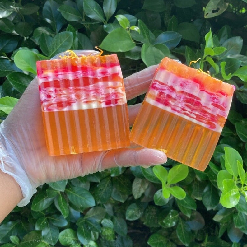 Puede incluir: Dos barras de jab&oacute;n hechas a mano con un dise&ntilde;o en capas. El jab&oacute;n es naranja, rosa y blanco. El jab&oacute;n est&aacute; en una mano que lleva un guante blanco.