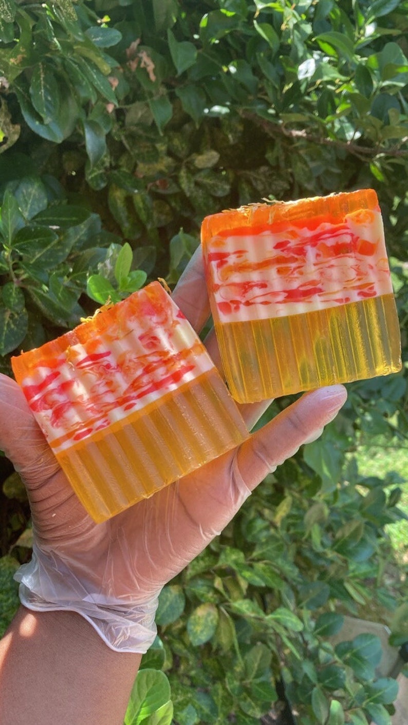 Puede incluir: Dos barras de jab&oacute;n hechas a mano con un dise&ntilde;o en capas. El jab&oacute;n es amarillo, naranja y blanco con un patr&oacute;n de remolino. El jab&oacute;n est&aacute; en una mano que lleva un guante de pl&aacute;stico transparente.