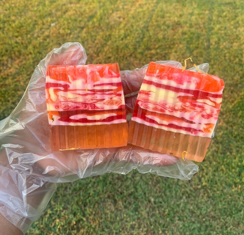 Puede incluir: Dos barras rectangulares de jab&oacute;n con un patr&oacute;n a rayas en tonos rojos, naranjas, blancos y amarillos. Las barras de jab&oacute;n est&aacute;n sobre un guante de pl&aacute;stico.