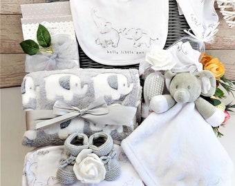 May include: A gray wicker basket filled with baby gifts, including a gray and white elephant print blanket, a white bib with an elephant design, a white hat, a gray elephant plush toy, a white blanket, and a pair of gray knitted booties. The basket is decorated with a white bow and a white rose.