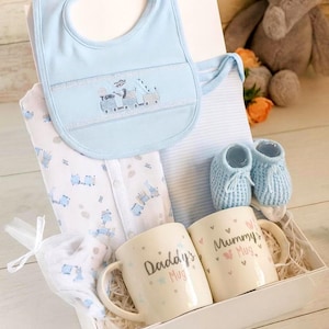 May include: A baby gift set in a white box. It includes a light blue bib with a train design, a onesie, two mugs that say "Daddy's Mug" and "Mummy's Mug", and blue baby booties. A small white bag with a baby item is also included.