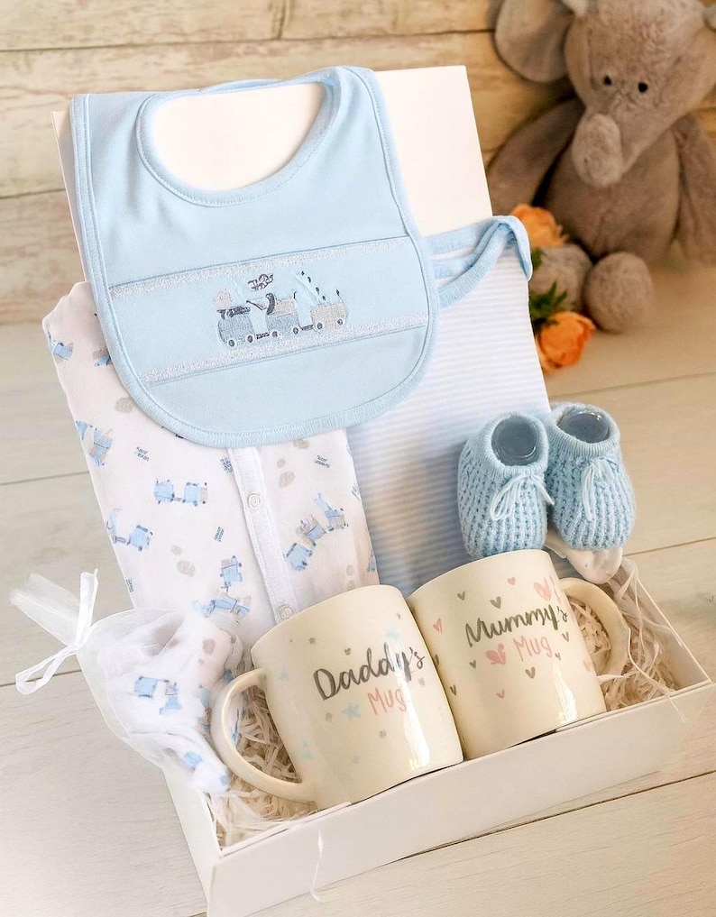 May include: A baby gift set in a white box. It includes a light blue bib with a train design, a onesie, two mugs that say "Daddy's Mug" and "Mummy's Mug", and blue baby booties. A small white bag with a baby item is also included.
