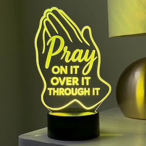 May include: A glowing LED night light with the words "Pray on it, over it, through it" in yellow script. The light is shaped like praying hands and sits on a black base. A gold lamp is in the background.