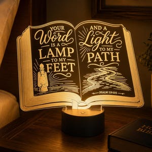 May include: An illuminated acrylic lamp shaped like an open book, featuring the engraved text 'YOUR WORD IS A LAMP TO MY FEET AND A LIGHT TO MY PATH' along with a lantern and path illustration. The lamp sits on a circular black base and glows warm.