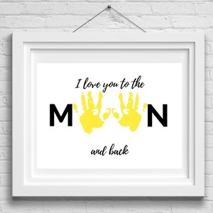 Pode incluir: Uma impressão emoldurada branca com o texto "I love you to the MOON and back" em preto. Duas impressões de mãos amarelas são usadas para formar o "OO" na palavra "MOON".