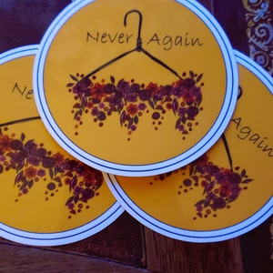 Op de afbeelding: Drie gele ronde stickers met een zwart hangertje silhouet en een bloemenkrans. De tekst "Never Again" staat op de bovenste sticker.