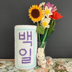 May include: A white cylinder with the Korean word "엄마" (meaning "mom") spelled out in purple beads. The cylinder is surrounded by a bouquet of artificial flowers, including sunflowers, white daisies, and pink and red roses. A small white bunny plush toy is sitting in front of the cylinder.