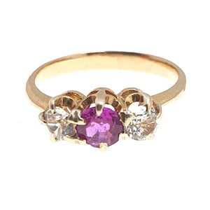 Może przedstawiać: Złoty pierścionek z centralnym, okrągłym, purpurowym kamieniem szlachetnym, otoczonym dwoma mniejszymi, przezroczystymi kamieniami. Pierścionek ma klasyczny wzór z trzema kamieniami.