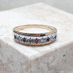 Pode incluir: Um anel de banda de ouro com uma fila central de pequenos rubis vermelhos redondos e diamantes brancos claros alternados, engastados num canal de tom prateado. O anel tem um acabamento em ouro polido nas bordas superior e inferior da banda.