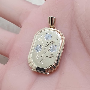 May include: An oval-shaped gold locket pendant featuring a scalloped edge and a central engraved floral design with three silver-toned flowers on stems. The locket includes a small gold bail at the top for attachment to a chain or necklace.