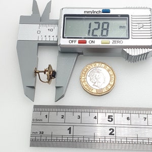 May include: A gold-tone earring with a black stone and a post back. The earring is being measured with a ruler and calipers. The ruler shows measurements in inches and millimetres. The calipers show a measurement of 12.8 mm.