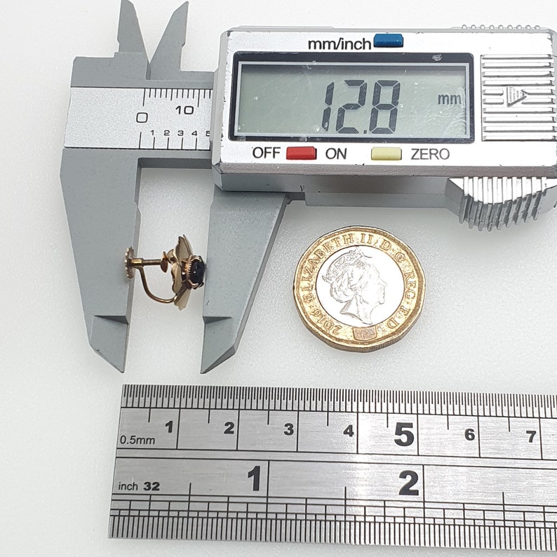 May include: A gold-tone earring with a black stone and a post back. The earring is being measured with a ruler and calipers. The ruler shows measurements in inches and millimetres. The calipers show a measurement of 12.8 mm.