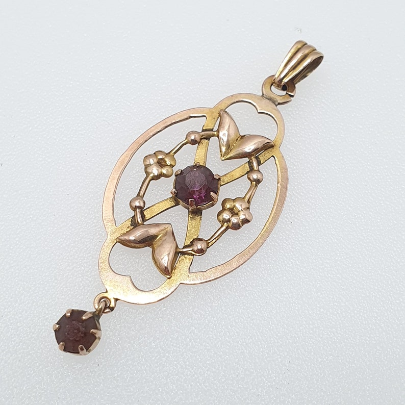 Pode incluir: Um pingente de ouro com um design delicado e intrincado que apresenta uma gema roxa central cercada por gemas menores. O pingente tem uma argola na parte superior para prender a uma corrente.