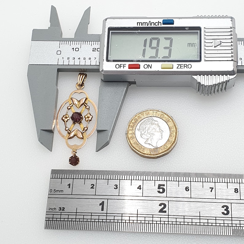 Pode incluir: Um pingente de ouro com um design delicado que apresenta uma pedra preciosa vermelha central e pedras preciosas menores ao redor. O pingente mede 1,93 polegadas de comprimento.