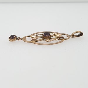 Pode incluir: Um pingente de ouro com um design delicado e intrincado com duas pedras preciosas de cor vermelho escuro. O pingente tem um la&ccedil;o na parte superior para prender a uma corrente.