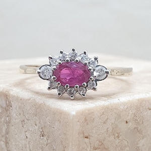May include: A gold ring featuring a central oval-cut ruby surrounded by a halo of small, round, clear gemstones. The ring's band is a simple, polished gold. The gemstones sparkle against the light, creating a classic and elegant design.