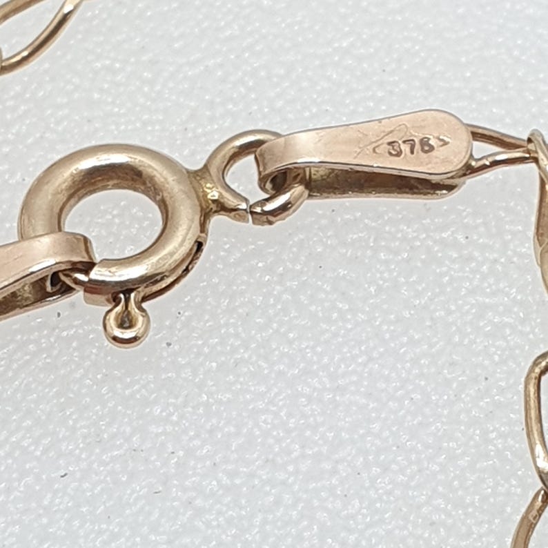 May include: Close-up of a gold-colored necklace clasp and chain. The clasp has a round ring and a hook closure. The clasp is stamped with the number "375". The chain links are small and delicate.