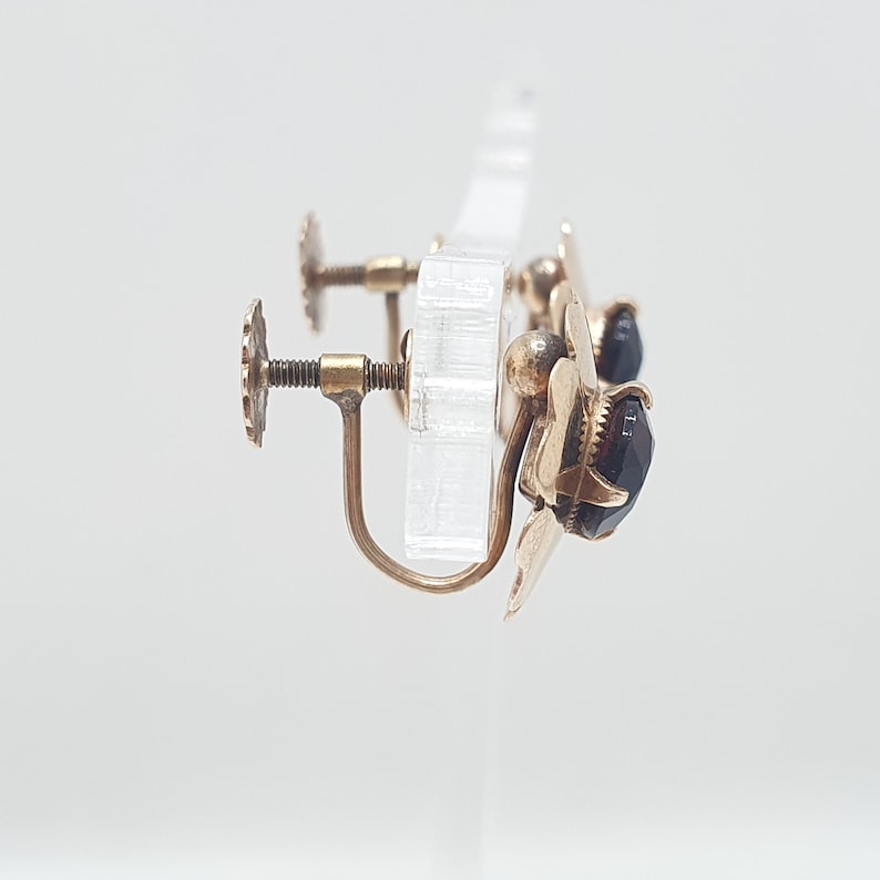 May include: A pair of gold-tone earrings with black gemstone accents. The earrings have a flower-shaped design and a screw-back fastening.