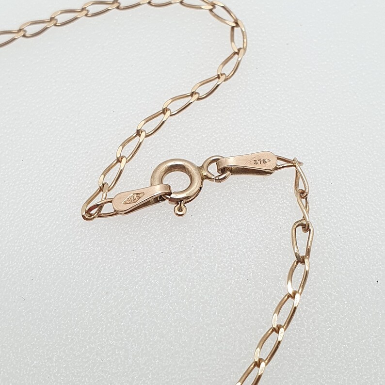May include: Close-up of a gold-colored necklace with an oval link chain. The clasp is visible, with a small loop and a connecting piece. The chain and clasp are on a light-colored surface.