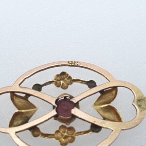 Pode incluir: Um pingente de ouro com uma gema vermelha no centro. O pingente tem a forma de um c&iacute;rculo com um design floral. A parte de tr&aacute;s do pingente &eacute; marcada com o n&uacute;mero "915".