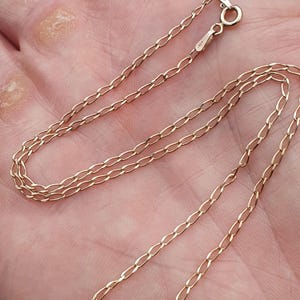 May include: A delicate gold-colored chain necklace with an oval link design. The necklace is shown against a skin-toned background, highlighting its fine details and the warm hue of the metal. The clasp is visible at the top.