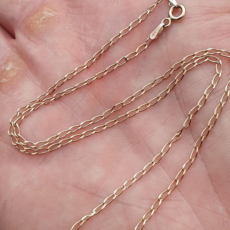 May include: A delicate gold-colored chain necklace with an oval link design. The necklace is shown against a skin-toned background, highlighting its fine details and the warm hue of the metal. The clasp is visible at the top.