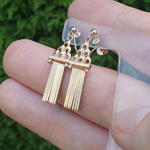 May include: A pair of gold-coloured earrings. Each earring has a decorative top with connected loops and a fringe of vertical bars. The earrings are held against a green background.