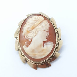 Puede incluir: Un broche de camafeo con un camafeo de concha tallada de perfil de mujer, engastado en un marco de metal dorado con un borde decorativo festoneado.