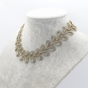 May include: A gold-toned necklace with a repeating leaf-like design. Each leaf shape has an openwork pattern and is accented with small, clear crystals. The necklace is displayed on a white mannequin bust.