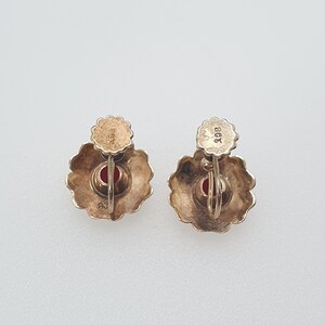 May include: A pair of gold earrings with red stones set in a flower design. The earrings are marked "9ct" on the back.