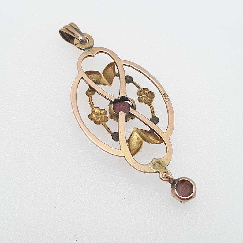 Pode incluir: Um pingente de ouro com uma gema vermelha no centro, rodeado por um delicado desenho floral. O pingente est&aacute; preso a uma corrente com uma pequena gema redonda na ponta.