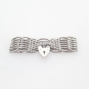 Puede incluir: Una pulsera plateada con un diseño tejido. La pulsera presenta un cierre de candado en forma de corazón en el centro. Las secciones tejidas están hechas de pequeños eslabones interconectados. La pulsera se muestra sobre un fondo blanco.