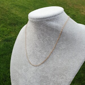 May include: A delicate gold-colored chain necklace displayed on a gray velvet bust. The chain has a fine, linked design and hangs gracefully. The background is a blurred view of green grass.