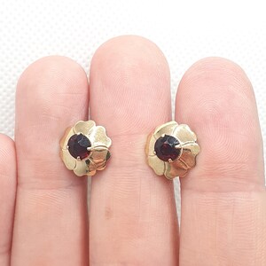 May include: A pair of gold earrings with a red gemstone in the centre of each earring. The earrings are shaped like a flower.