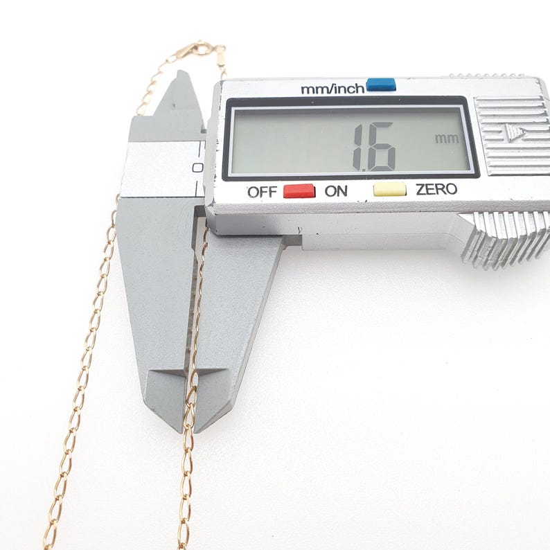 May include: A gold-colored chain necklace is measured with a digital caliper. The digital display reads 1.6 mm. The caliper is silver with black text and red and blue accents. The chain is thin and delicate.